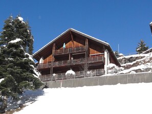 A wooden chalet with balconies, surrounded by snow-covered trees and a clear blue sky.