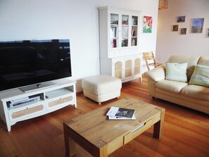 Ett vardagsrum med en platt-TV, en vit soffa, ett träbord och ett vitt skåp med glasdörrar.