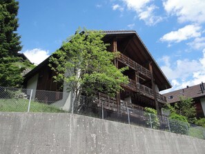 Un bâtiment traditionnel en bois, doté d’un balcon, entouré d’une clôture en treillis métallique et de verdure.