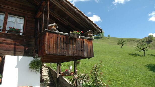 A wooden house with a balcony, a white wall, and a green hill in the background. - Serene Farmhouse in Habkern with Parking (Habkern)
