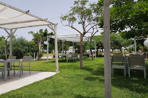 Un área de estar al aire libre con mesas y sillas bajo una pérgola blanca, rodeada de césped y árboles.