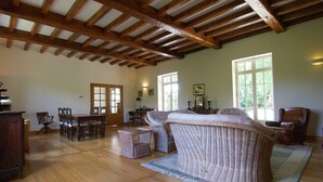 A spacious room with wooden beams on the ceiling, a dining area with chairs and a table, and a wicker sofa.