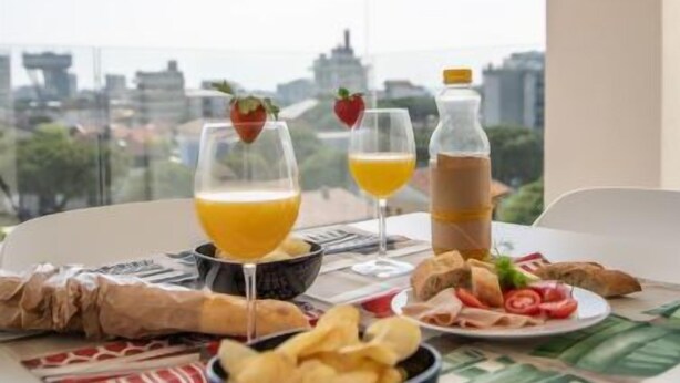 En balkon med et bord dækket til morgenmad, inklusive en skål chips, en tallerken med skivede tomater og grønt, en juiceflaske og to glas appelsinjuice med jordbær.
