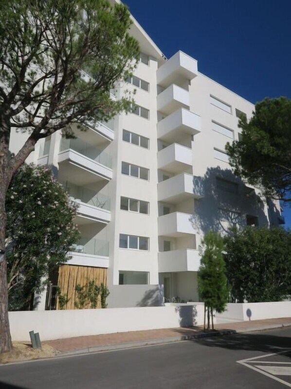 A multi-story white building with balconies, surrounded by trees and a clear sky.