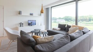 A modern living room with a large grey sofa, a white chair, a small coffee table, and a television on a white shelf.