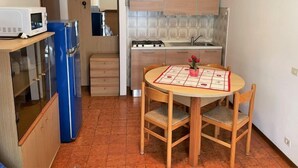 Cozinha compacta com micro-ondas, geladeira, fogão e armários de madeira. Há uma pequena mesa de jantar com cadeiras e um vaso de flores.