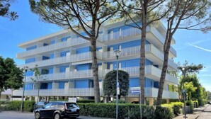Um edifício com vários andares, varandas, área de estacionamento e um carro estacionado em frente.