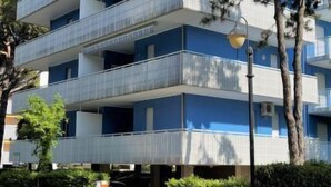 Um edifício residencial com várias andares, varandas, céu claro e vegetação ao redor.