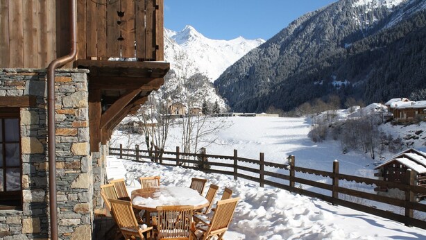 A wooden cabin with stone walls, a wooden deck, and a round table with chairs in a snowy landscape with mountains in the background. - Luxury flat with sauna in the French Alps (Champagny en Vanoise)