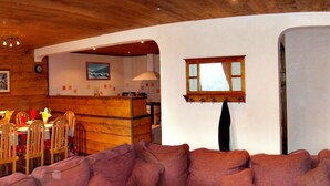 A cozy living room with a sofa, wooden dining table, chairs, and a kitchen area with a window and a picture on the wall. - Luxury flat with sauna in the French Alps (Champagny en Vanoise)