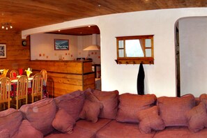 Een gezellige woonkamer met een bank, een houten eettafel, stoelen en een keukenhoek met een raam.