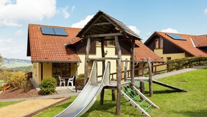Un terrain de jeux équipé d’un toboggan et d’une structure d’escalade, situé devant un bâtiment dont le toit est recouvert de tuiles rouges et la façade est jaune.