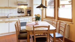 Une cuisine avec des meubles blancs, une table à manger en bois avec des chaises, un luminaire suspendu et une fenêtre ornée de rideaux en dentelle.