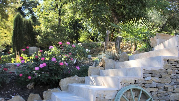 A stone staircase leading up to a stone wall with a wheel, surrounded by lush greenery and pink flowers. - Modern gîte with pool on large property of owner, 1 km from Provençal village (Faucon)