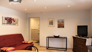 A living room with a red sofa, a wooden dining table, a TV on a wooden cabinet, and artwork on the walls. - Modern gîte with pool on large property of owner, 1 km from Provençal village (Faucon)