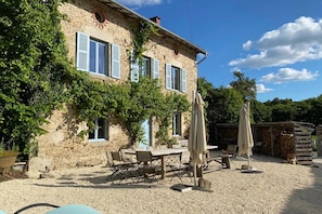 Ein Steinhaus mit blauen Fensterläden, einer Schotterterrasse, Gartenmöbeln und einer Pergola.