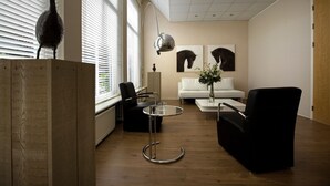 A modern living room with a wooden floor, a black leather sofa, a white armchair, a glass coffee table, and a black leather chair. There is a wooden podium with a decorative item on top, a floor lamp, and two framed artworks on the wall.