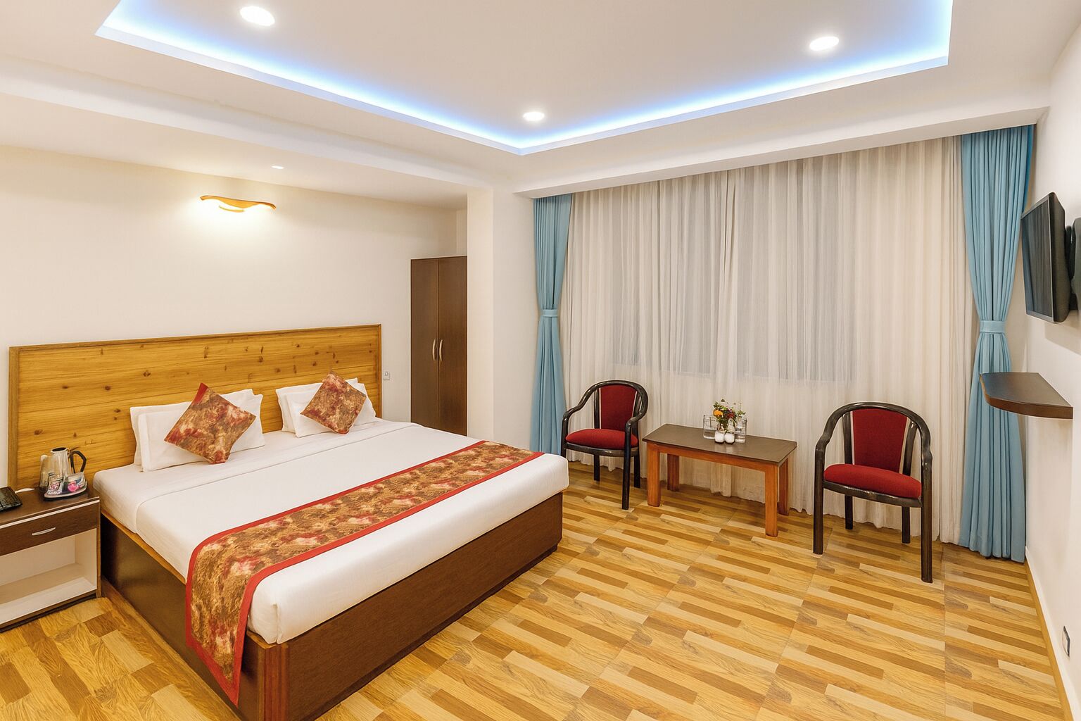 A hotel room with a wooden bed, a desk, two chairs, a TV, and a window with curtains.