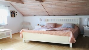 Ein Schlafzimmer mit Holzboden, einem weißen Bett mit rosa Bettwäsche, einem Nachttisch mit Lampe und einem kleinen Fenster.