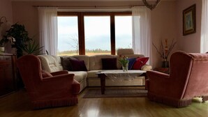 Ein Wohnzimmer mit einem beigefarbenen Sofa, zwei roten Sesseln, einem Holztisch und Blick ins Freie durch große Fenster.