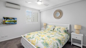 A bedroom with a bed, two bedside tables, a ceiling fan, an air conditioner, a wall-mounted TV, a window with blinds, and a decorative mirror. - Water Views with a Splash of Luxury (BONGAREE)