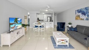 A modern living room with a TV, white furniture, a grey sofa, and a kitchen in the background. - Water Views with a Splash of Luxury (BONGAREE)