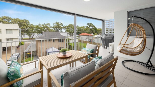A balcony with a wooden table, cushioned seating, a hanging chair, and a view of neighboring buildings and trees. - Water Views with a Splash of Luxury (BONGAREE)