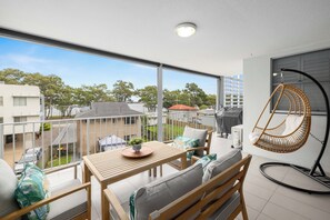 A balcony with a wooden table, cushioned seating, a hanging chair, and a view of neighboring buildings and trees.