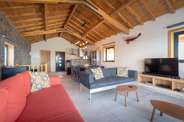 Un salón espacioso con un alto techo de madera, un sofá gris con cojines estampados, un sofá rojo, una mesa de centro de madera y un televisor sobre un mueble de madera.