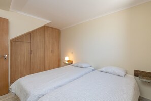 A bedroom with two beds, a wooden wardrobe, a bedside table with a lamp, and a wall-mounted shelf.