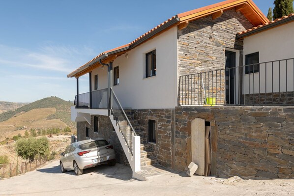 Una casa de dos pisos con fachada de piedra, un balcón y un coche estacionado frente a ella.