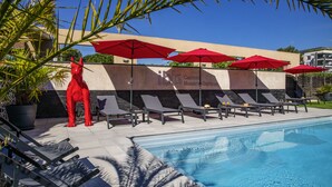 A pool area with lounge chairs and red umbrellas, a red statue, and a wall with the text "ibis Cannes Moujoux". - Hôtel Ibis Cannes Mouans Sartoux (Mouans-Sartoux)