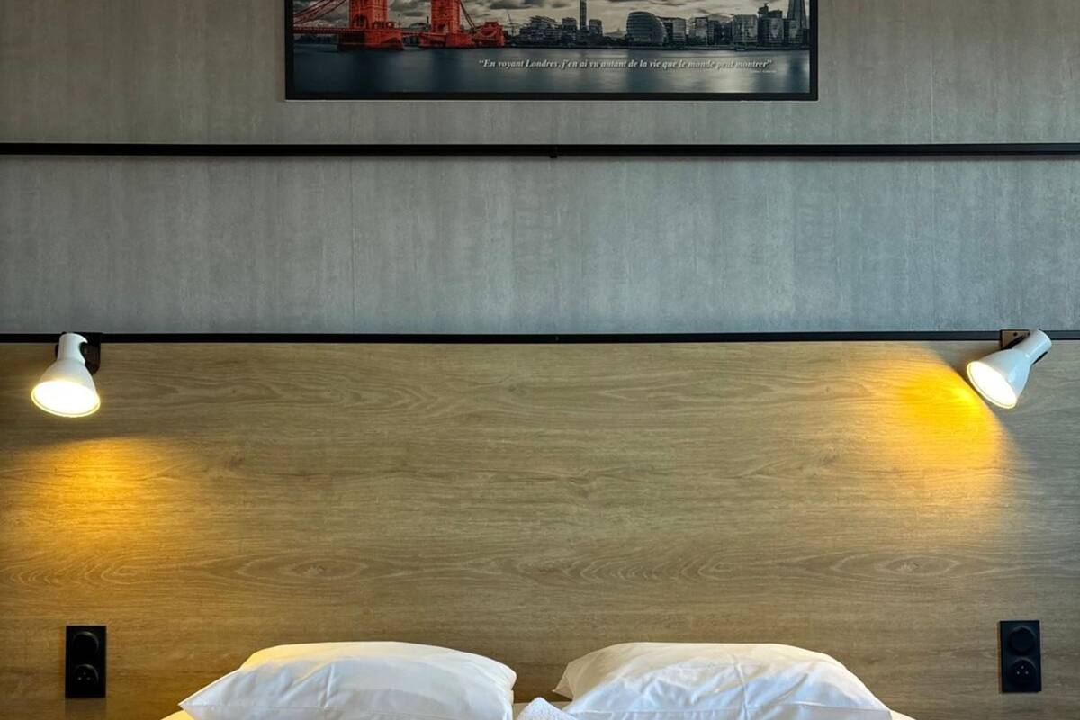 A hotel room with a bed, two bedside lamps, a painting of a bridge, and a rolled towel on the bed.