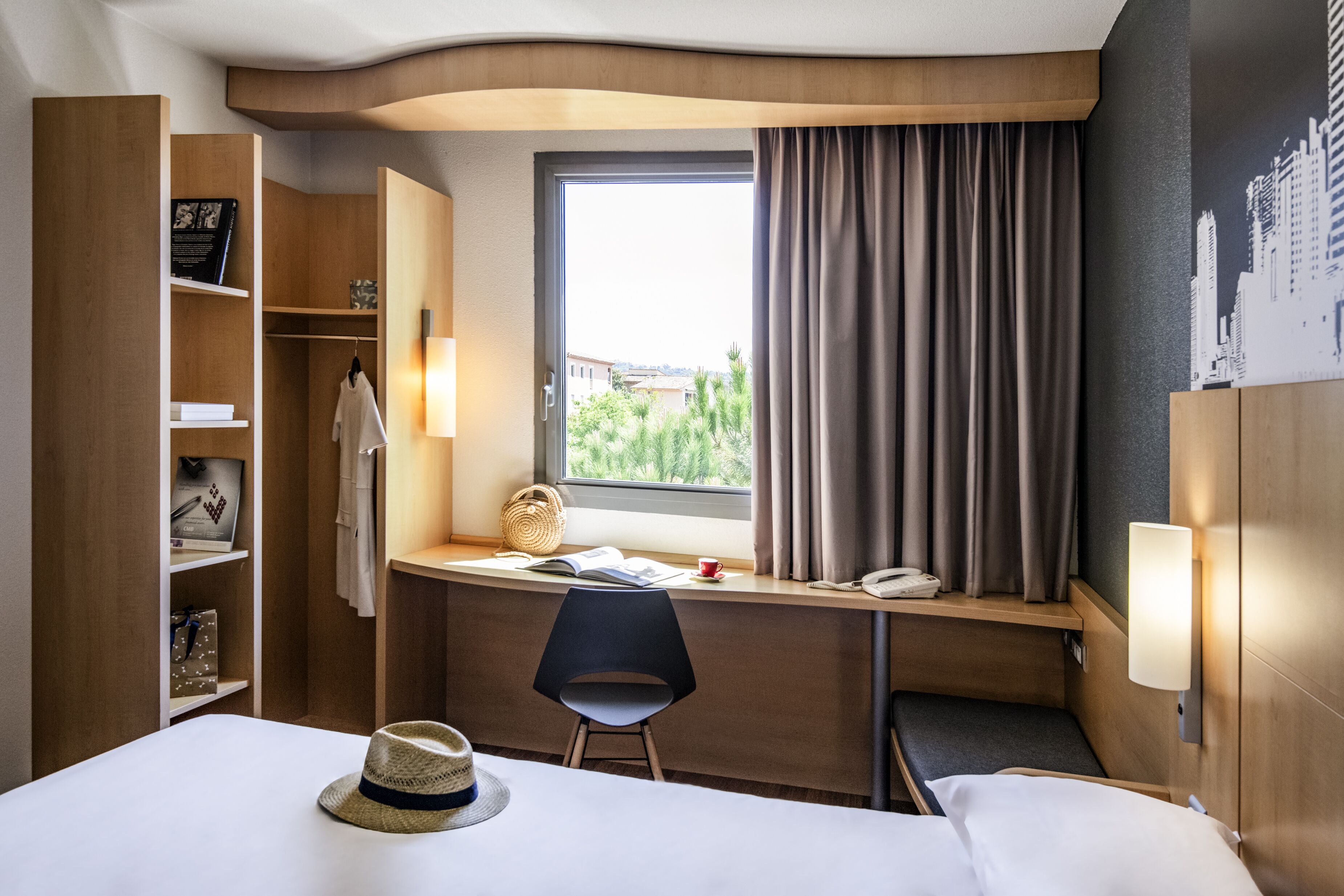 Quarto de hotel com cama, secretária com cadeira, guarda-roupa, janela com cortinas e vista para a cidade.