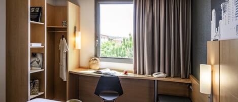 Quarto de hotel com cama, secretária com cadeira, guarda-roupa, janela com cortinas e vista para a cidade.