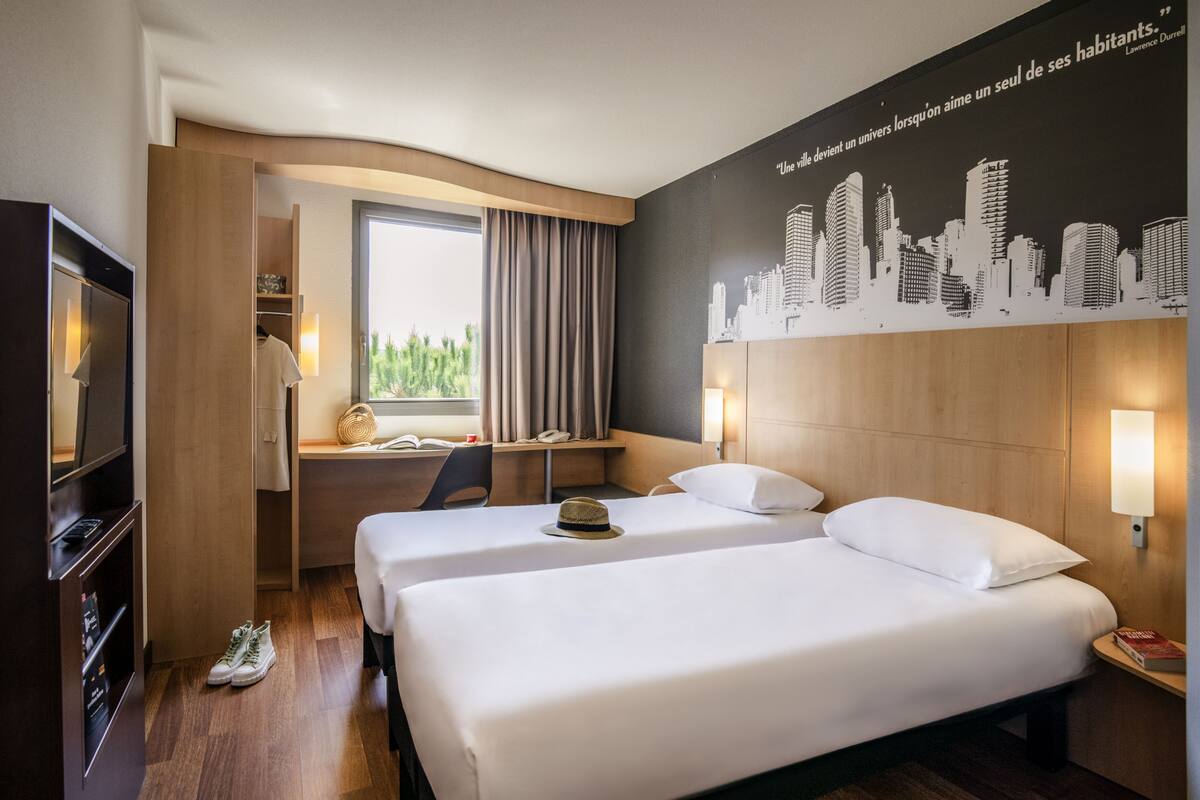 A hotel room with two beds, a desk, a TV, and a cityscape mural on the wall.