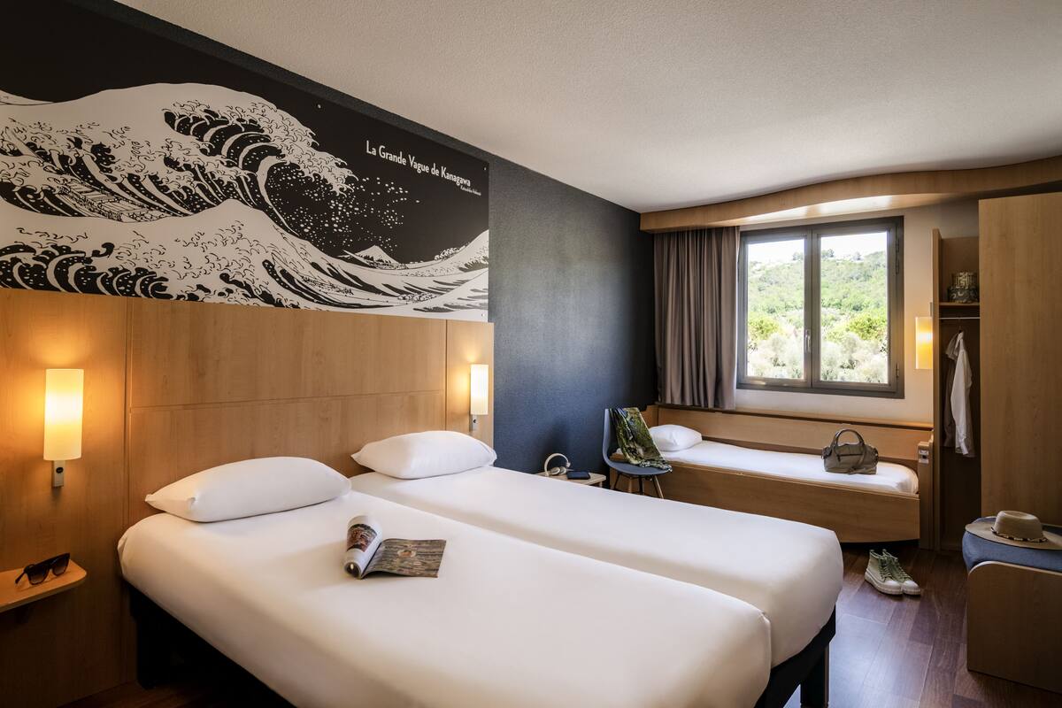 A hotel room with two beds, a large wave mural, a window with a view, and a wardrobe.