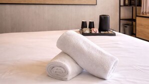 A neatly made bed with a rolled towel, a flat-screen TV mounted on the wall, a shelf with toiletries, and a trash bin. - Hôtel Ibis Cannes Mouans Sartoux (Mouans-Sartoux)