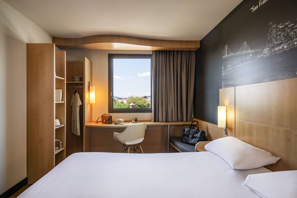 A hotel room with a bed, a desk, a chair, a wardrobe, and a view of the city.