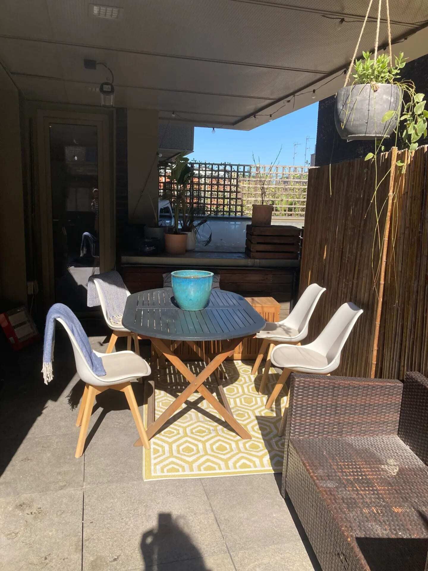 An outdoor patio with a round table and four chairs, a hanging plant, and a wooden fence in the background.