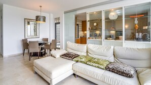 Una sala moderna con un sofá seccional, un comedor con mesa y sillas, y una cocina con armarios de vidrio e iluminación colgante.