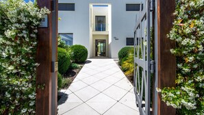 A modern building entrance with a clear pathway leading to a door, surrounded by greenery and a wooden gate with a number sign.