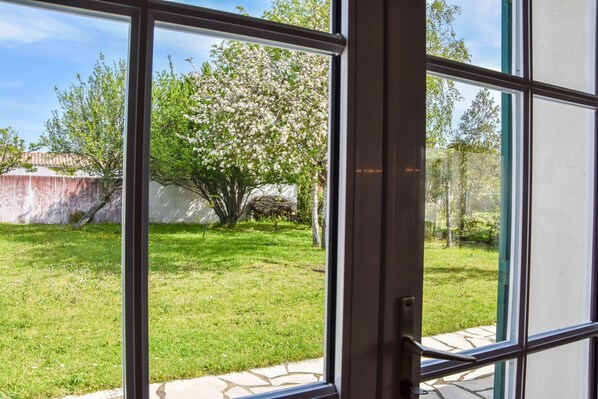 A view through a window with a stone pathway leading to a grassy yard with trees and a white wall.