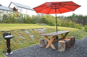 Een houten picknicktafel met een rood parasol, omringd door houten krukjes, op een grindpad met een tuin en een huis op de achtergrond.