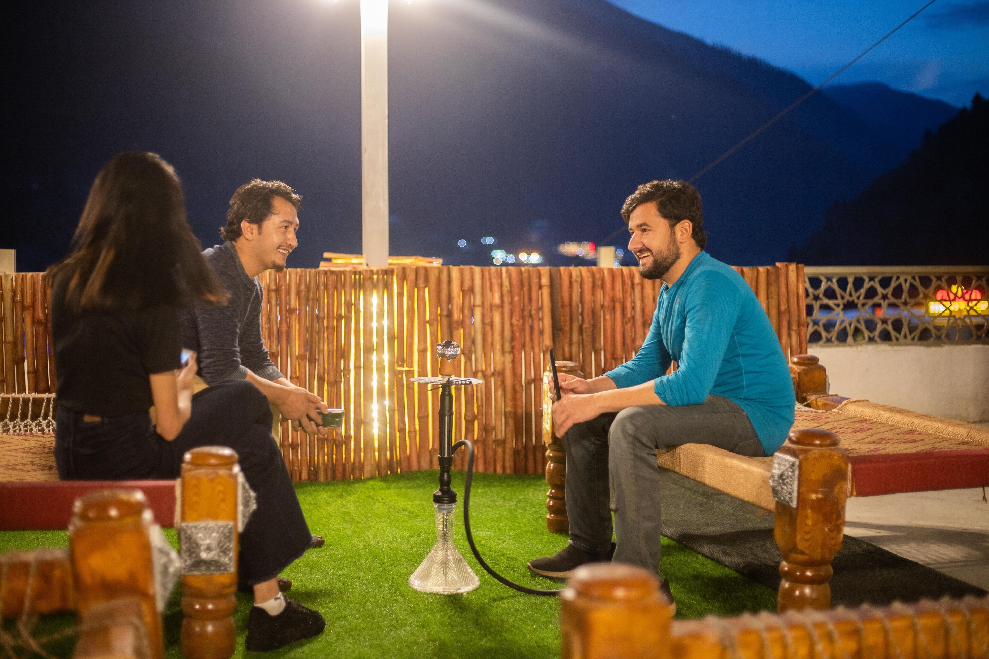 Three people seated on a bench outdoors at night with a hookah in the center, surrounded by wooden furniture and a bamboo fence.