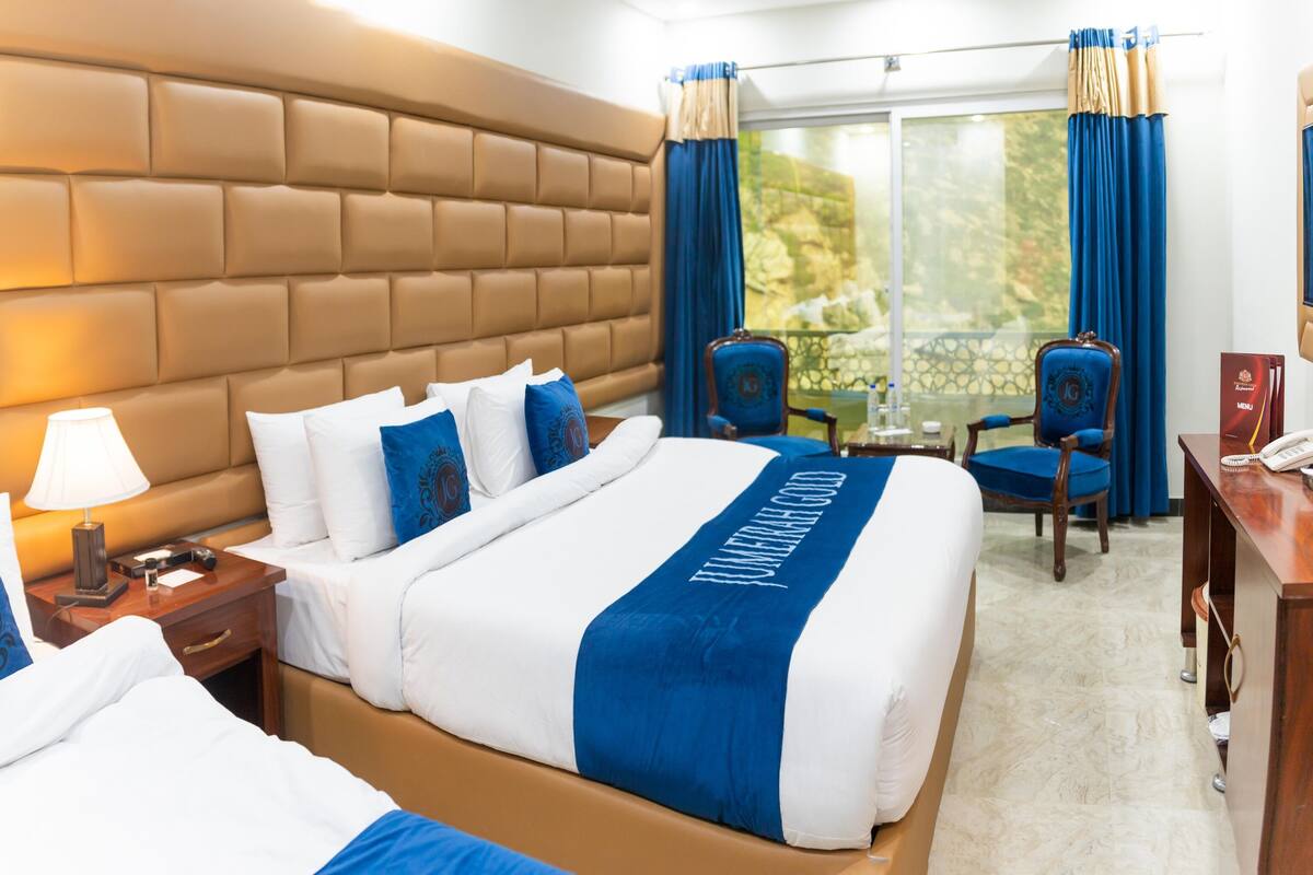 A hotel room with a large bed, a wooden headboard, a desk with a lamp, and a view of the outdoors through a window with curtains.