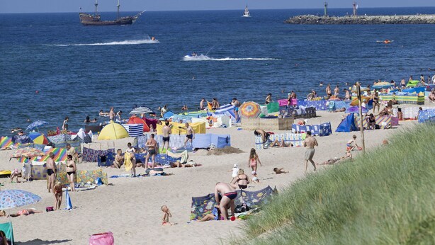 Ein voller Strand mit Menschen, die sonnenbaden und schwimmen, Zelten und einem Schiff im Hintergrund.