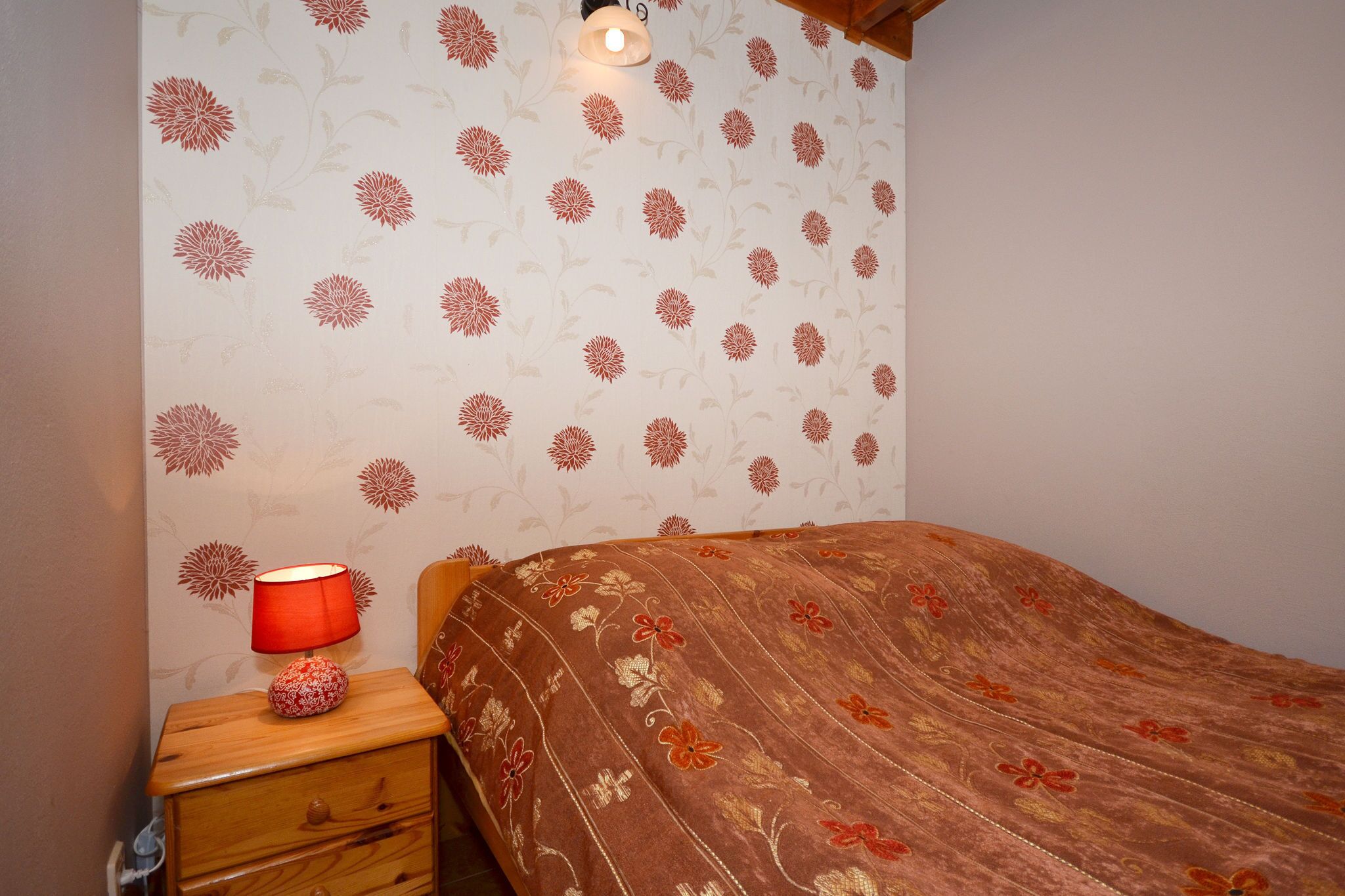 A bedroom with a floral wallpaper, a wooden nightstand with a red lamp, and a bed with a floral bedspread.