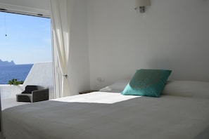 A bedroom with a bed, a chair, a window with curtains, and a view of the sea.