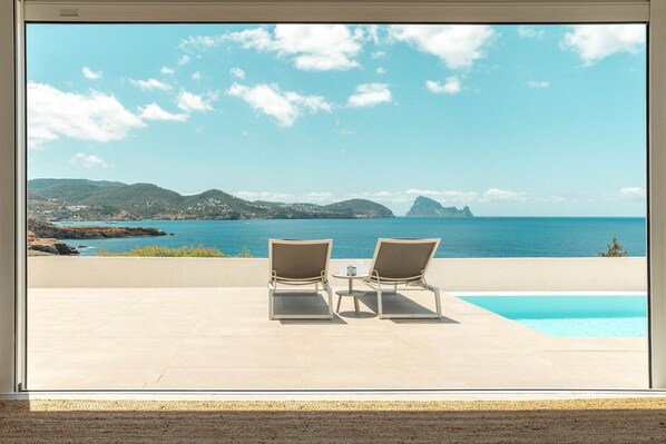 A terrace with two lounge chairs and a small table, overlooking a pool and a coastal landscape.
